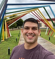 Foto de Fabricio Paiva Mota: selfie de um homem branco, de cabelo castanho, curto, sem barba, sorrindo. No fundo, uma praça com uma instalação artística de madeira em cores.