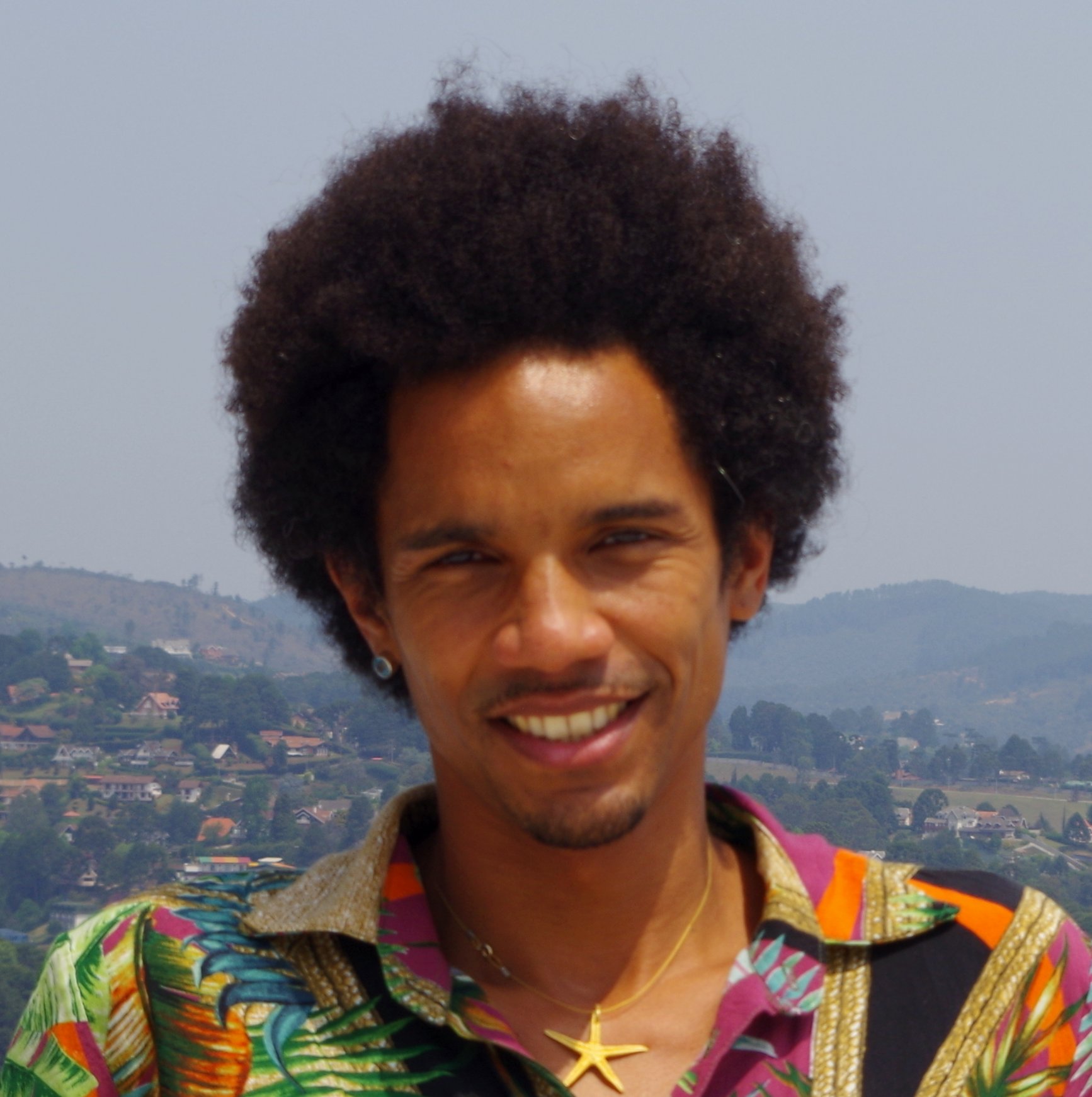 Foto de Alexander Yao Cobbinah: homem negro, jovem, de cabelo crespo e alto, cavanhaque bastante curto no queixo, sorridente. Veste uma camisa com muitas cores e um colar um pingente de tamanho médio, em formato de estrela, na cor amarela. No fundo, uma paisagem de montanhas distante.
