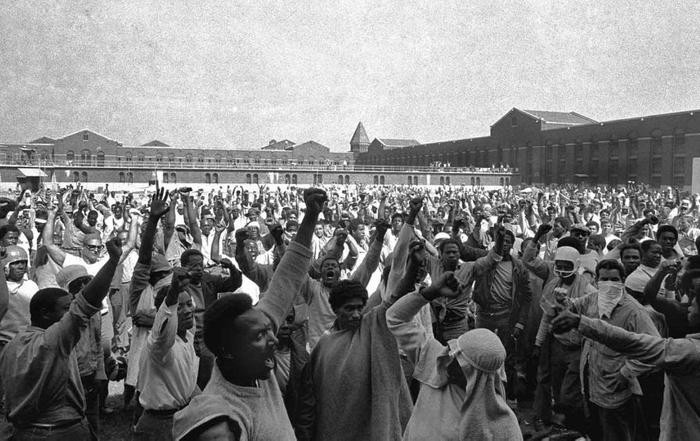 Black and white image of a crowd of Black inmates with raised fists during the 1971 Attica Prison Uprising.