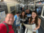 Group of five smiling on a NYC subway. One holds a map. Posters and seats in background. Casual attire, colorful shirts. Urban exploration mood. Take a private neighborhood walking tour of New York City with Adam Guy of I Know A Guy NYC Tours.