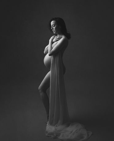 Mulher grávida em uma sessão fotográfica. Ela está em pé, de perfil, sem roupa, segurando um pano que vai até o chão na lateral do corpo. Os braços cruzados na frente dos seios. A foto está em preto e branco.