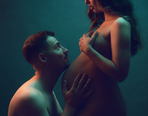 Casal em sessão fotográfica de grávida, com homem com mãos na barriga da mulher, olhando para ela e com queixo encostado à barriga. A fotografia tem vários tons de cor misturando-se.