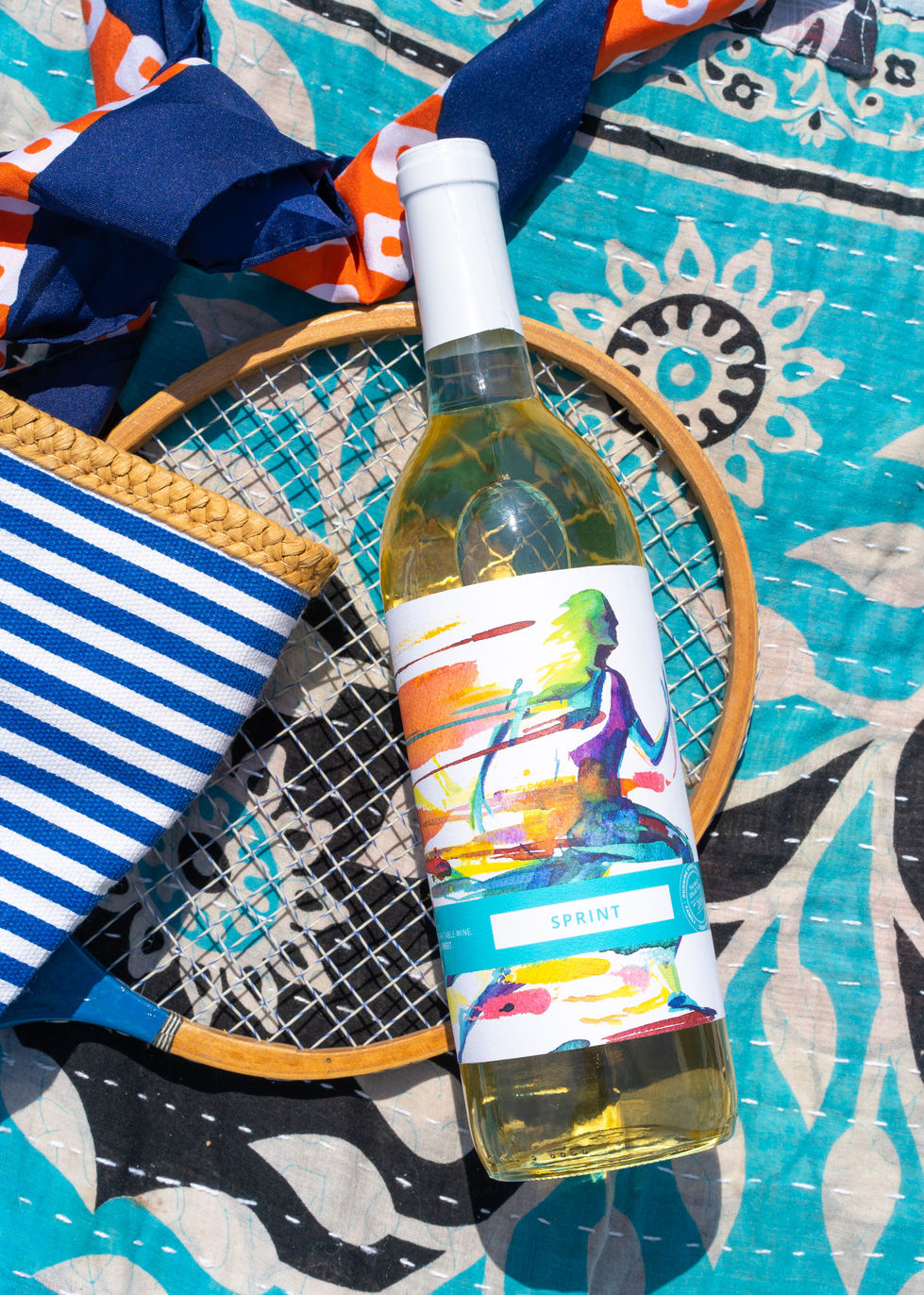 Bottle of Sprint wine by Running Vines winery resting on a tennis racket and a blue tribal quilt