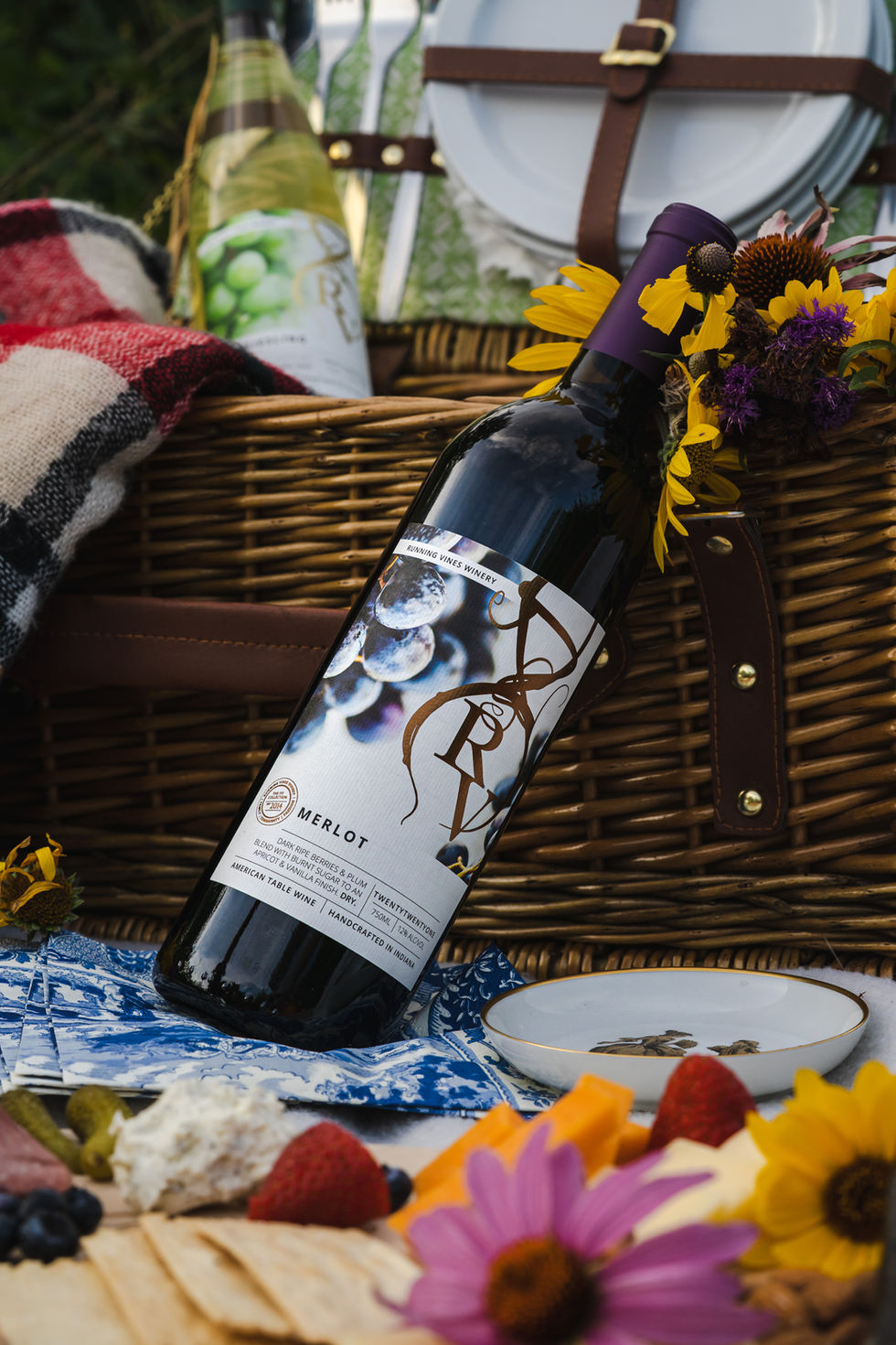 Bottle of Running Vines Merlot leaning against a whicker picnic basket with a charcuterie tray in the foreground