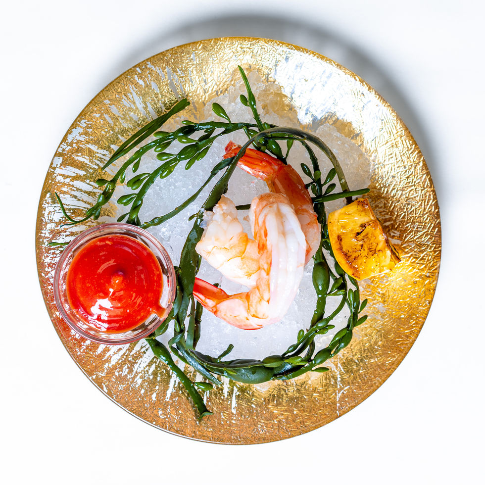 Gold gilt plate featuring two shrimp and small container of cocktail sauce on a bed of crushed ice
