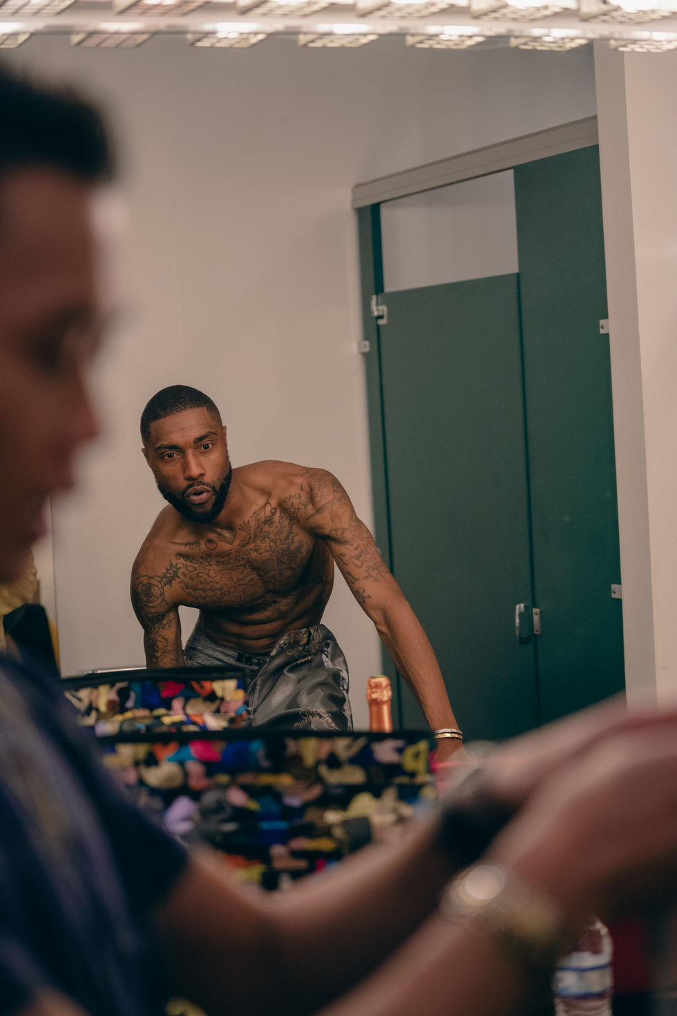 Shirtless Youth Soul Love getting ready backstage as clothing designer Nicolas Nguyen looks through a clothing rack in the foreground