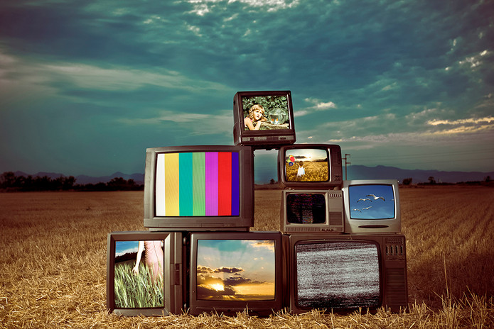 Pirámide de televisores antiguos apilados en un campo de trigo dorado bajo un cielo dramático