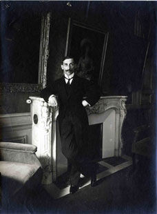 Man in a suit leans casually on an ornate fireplace in a dimly lit, elegant room. A painting and plush chairs are visible in the background.