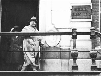 Man in a trench coat and hat exits a building, stepping down stairs. There's a sign "Goudstikker," and the setting is urban and historic.