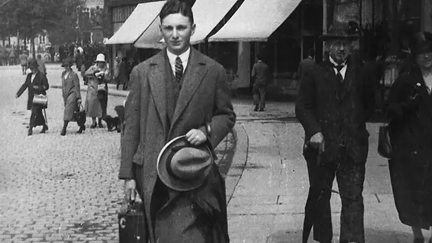 A man in a suit and tie walks on a cobblestone street holding a hat. People with hats and coats stroll behind him. Storefronts are visible.