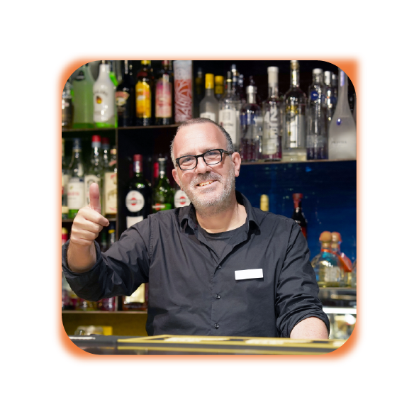 Smiling bartender giving a thumbs-up behind a bar counter with bottles of liquor in the background