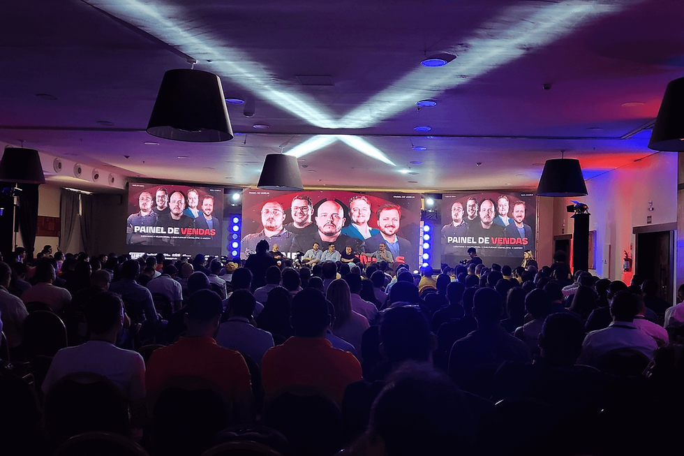 Fotografia em plano aberto e contraluz de um auditório lotado durante um evento corporativo. Ao fundo, um palco com painel de LED triplo exibe a identidade visual do "Painel de Vendas" com fotos dos palestrantes. O teto do espaço é iluminado por feixes de luz branca que se cruzam em formato de "X", contrastando com a plateia que aparece em silhueta.