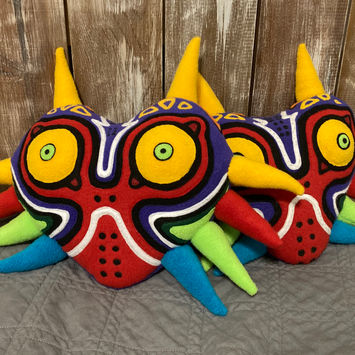 A photograph of two identical fleece plushies. They are multicolored masks, primarily red and purple with back and white striping and large yellow eyes. The masks have 10 large multicolored horns, 2 atop the head and 8 along the cheeks