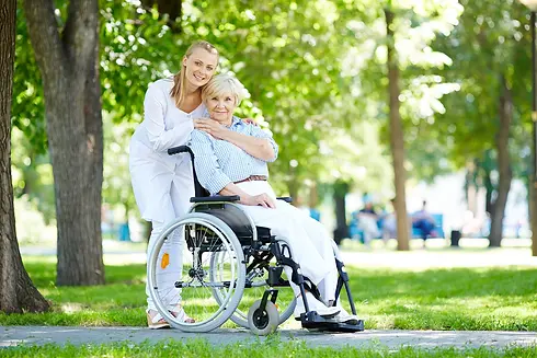 Eine Ältere Dame sitzt im Rollstuhl in einem Stadtpark und wird von Ihrer Alltagsbegleiterin liebevoll umsorgt.