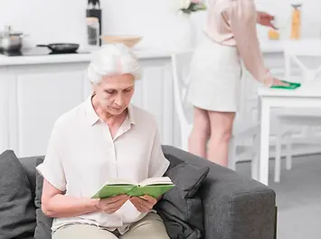 Eine Seniorin sitzt auf der Couch und liest ein Buch, während ihre Haushaltshilfe den Küchentisch abwischt