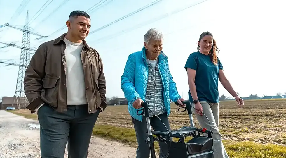 Eine Seniorin unternimmt in Begleitung ihrer Alltagshelfer einen Spaziergang bei schönem Wetter.