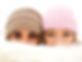 Portrait of two girls peeking from behind a fuzzy white blanket, wearing beanies (one brown and one pink), with only their eyes visible. The focus is on their curious and playful expressions