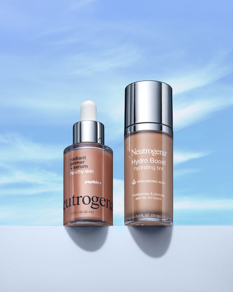A beautifully lit product shot featuring Neutrogena’s Radiant Primer + Serum with peptides and Hydro Boost Hydrating Tint with hyaluronic acid, set against a serene blue sky background. The sleek glass bottles and reflective silver caps highlight the skincare-focused formulas designed to hydrate, plump, and enhance healthy-looking skin.