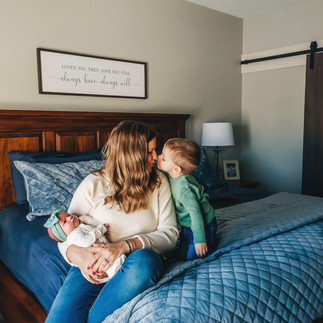 mom holding newborn on blue bed and kissing toddler