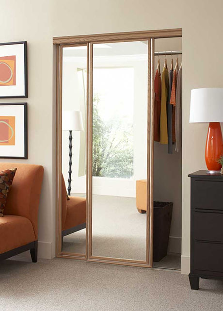 Mirrored sliding closet doors with clothes hanging inside, a lamp and a small ottoman visible in the reflection.