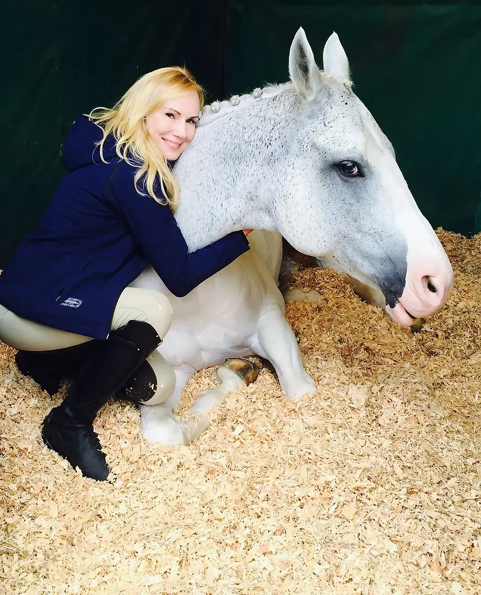 Blonde woman embracing a white horse while sitting on the ground