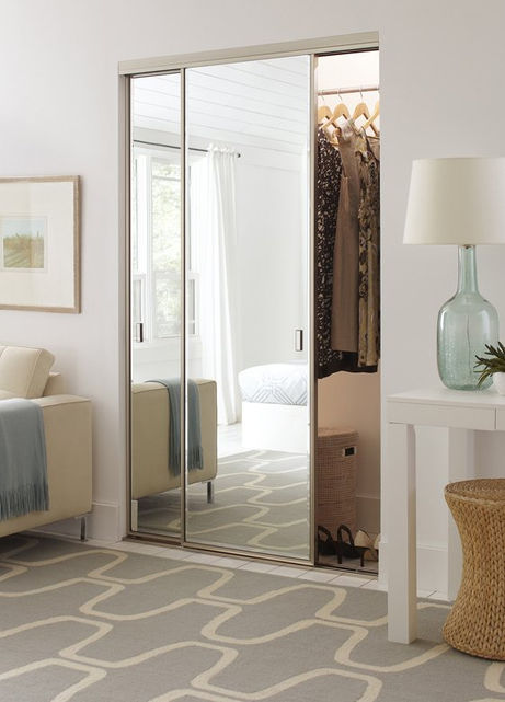 Mirrored closet doors with clothes hanging inside, a view of a bedroom with a bed, a couch and a lamp in the room