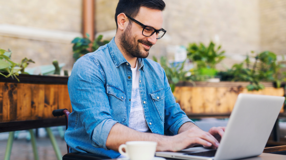 Man in blue denim shirt smiling, typing on laptop at outdoor cafe with plants and brick wall in background, cup of coffee on table.