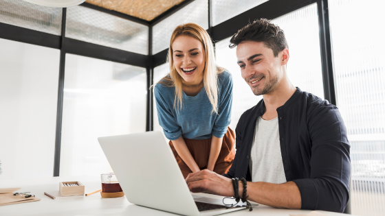 Two people smiling and looking at a laptop in a modern office. A woman in blue leans over, and a man types.