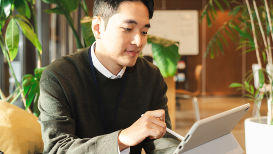 Man in a green sweater using a stylus on a tablet in a bright room with plants. He appears focused.