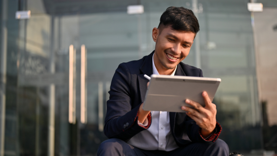 Smiling man in suit uses a tablet outside a glass building. Daylight, professional setting, focused and positive mood.