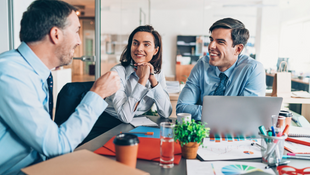 Three business leaders in office attire converse cheerfully around a table with laptops and documents.