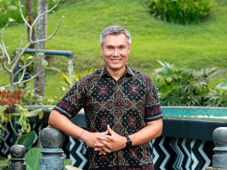 Smiling man in patterned shirt stands in a lush garden, with green grass and pool in the background, hands clasped in front.