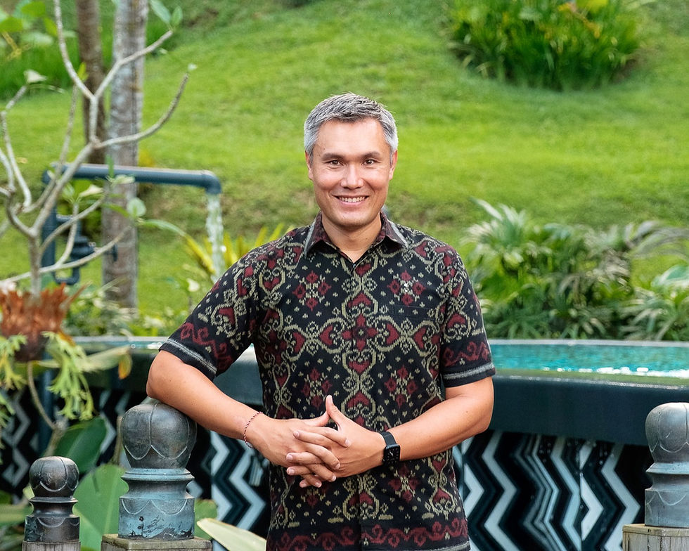 Smiling man in patterned shirt stands in a lush garden, with green grass and pool in the background, hands clasped in front.