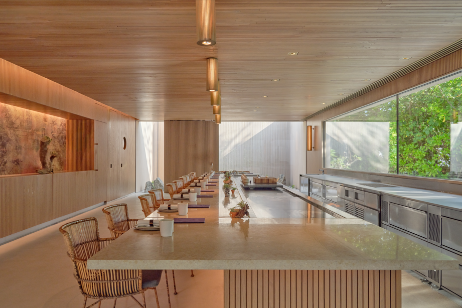 Modern, wooden dining area with a long table and wicker chairs. Large windows showing greenery. Warm lighting creates a cosy atmosphere.