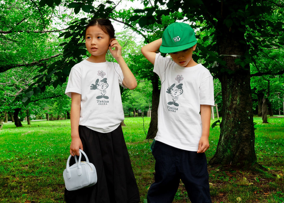 Two children in white "Patina Osaka" shirts stand in a green park. One holds a small bag; both adjust earbuds, appearing thoughtful.