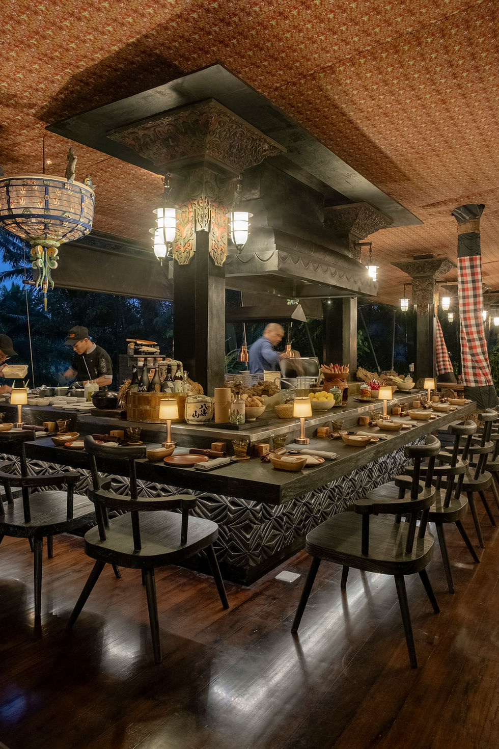 Cooks prepare food at a decorative, rustic open-kitchen restaurant. Dim lighting, patterned ceiling, and set tables create a cosy atmosphere.