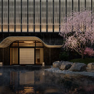 Elegant building entrance with a curved roof, cherry blossom tree, rocks, and a misty pond in the foreground. Calm and serene atmosphere.