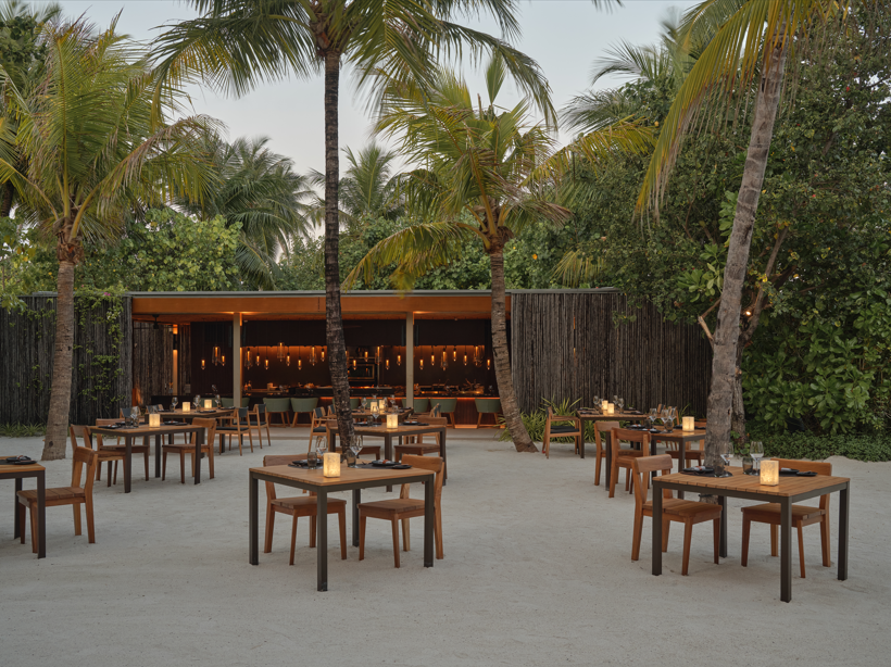 Outdoor restaurant scene with wooden tables under palm trees, candles on each table. Lush greenery surrounds the open-air setting. Tranquil mood.