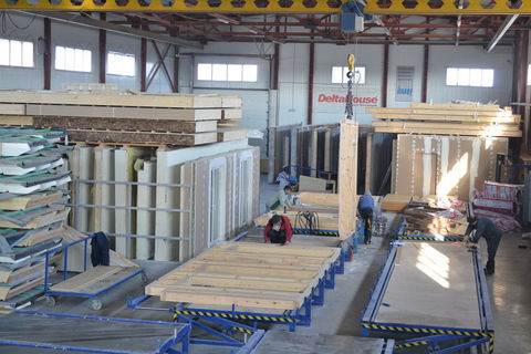 Wall panels and wood components on a factory assembly line with crane systems and workers.