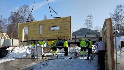 Construction team guiding a crane-lifted prefabricated wall module into place on a snowy modular building site.