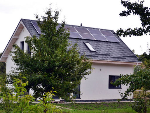 Modern white house with solar panels on a gray roof, surrounded by green trees and grass. Overcast sky in the background.