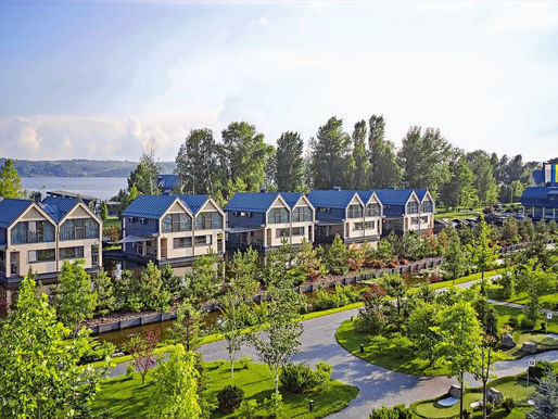 Modern houses with blue roofs line a lush green street, bordered by trees. A serene lake and blue sky form the backdrop, creating a tranquil scene.