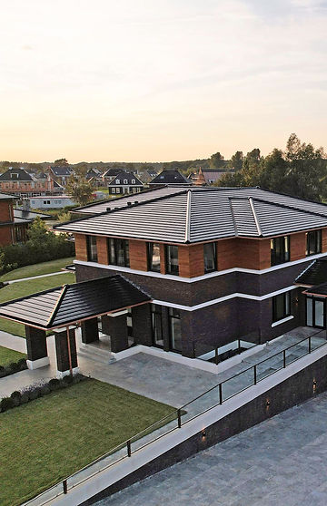 Modernes zweistöckiges Haus mit dunkler Klinkerfassade, großen Fenstern und großzügigem Garten, im Hintergrund ein Wohngebiet.
