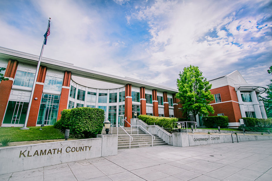 The Klamath County Government Center with large windows and masonry brick siding, with landscaped bushes and trees.