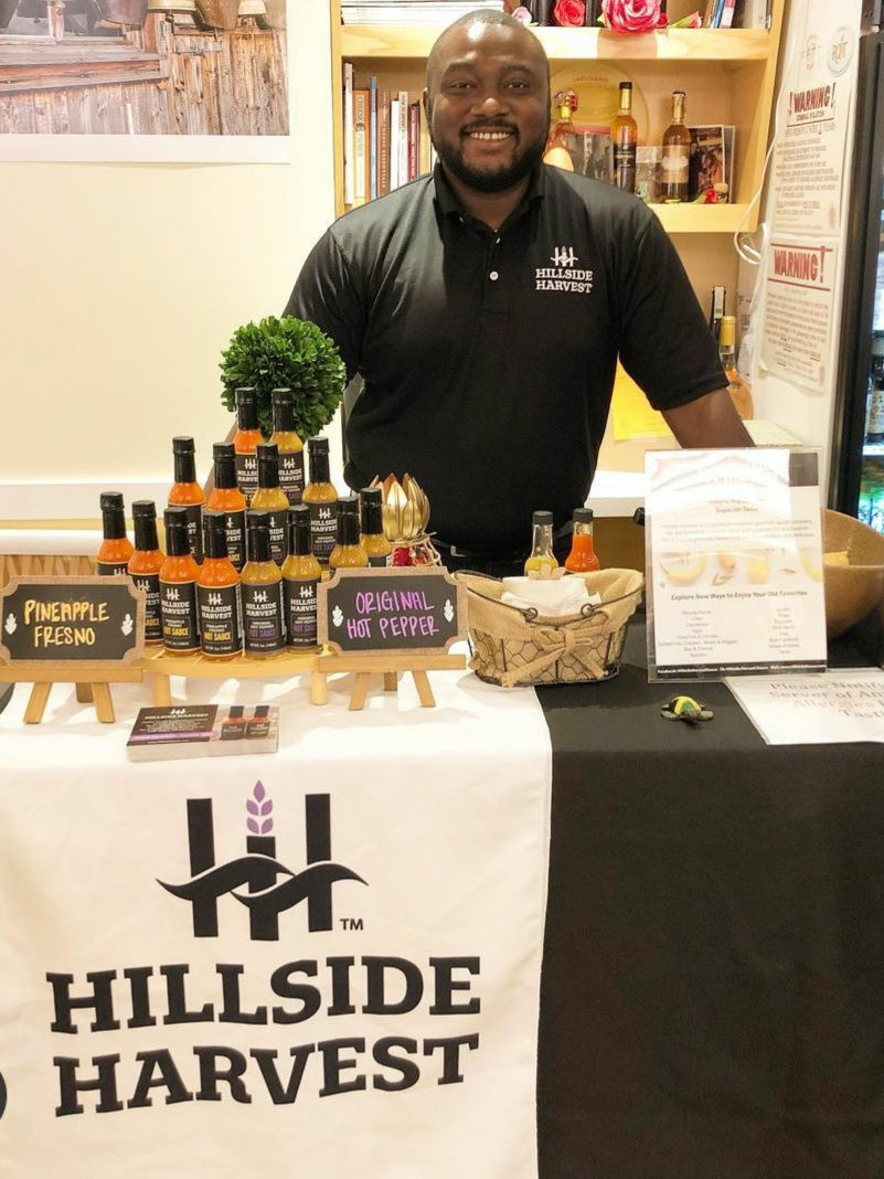Smiling man stands behind a display of hot sauces labeled "Pineapple Fresno" and "Original Hot Pepper." The tablecloth reads "Hillside Harvest."