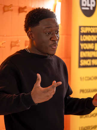 "A man in a black jumper gestures expressively while speaking at the Incubator Plus Programme Introductory Session. Behind him is an orange wall and a Business Launchpad banner promoting support for young entrepreneurs.