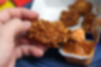 Hand holding crispy fried chicken drumstick dipped in orange sauce, over a dark blue plate. Blurred chicken pieces in background.