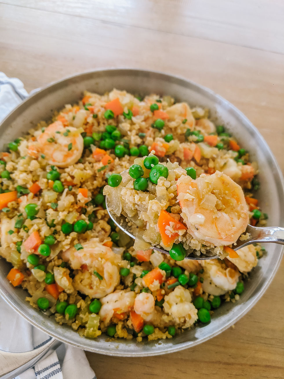 A close up of a stainless steel pan, with shrimp fried cauliflower rice is shown. A spoon is taking a large scoop to show a close up of a piece of shrimp and the rice mixture.