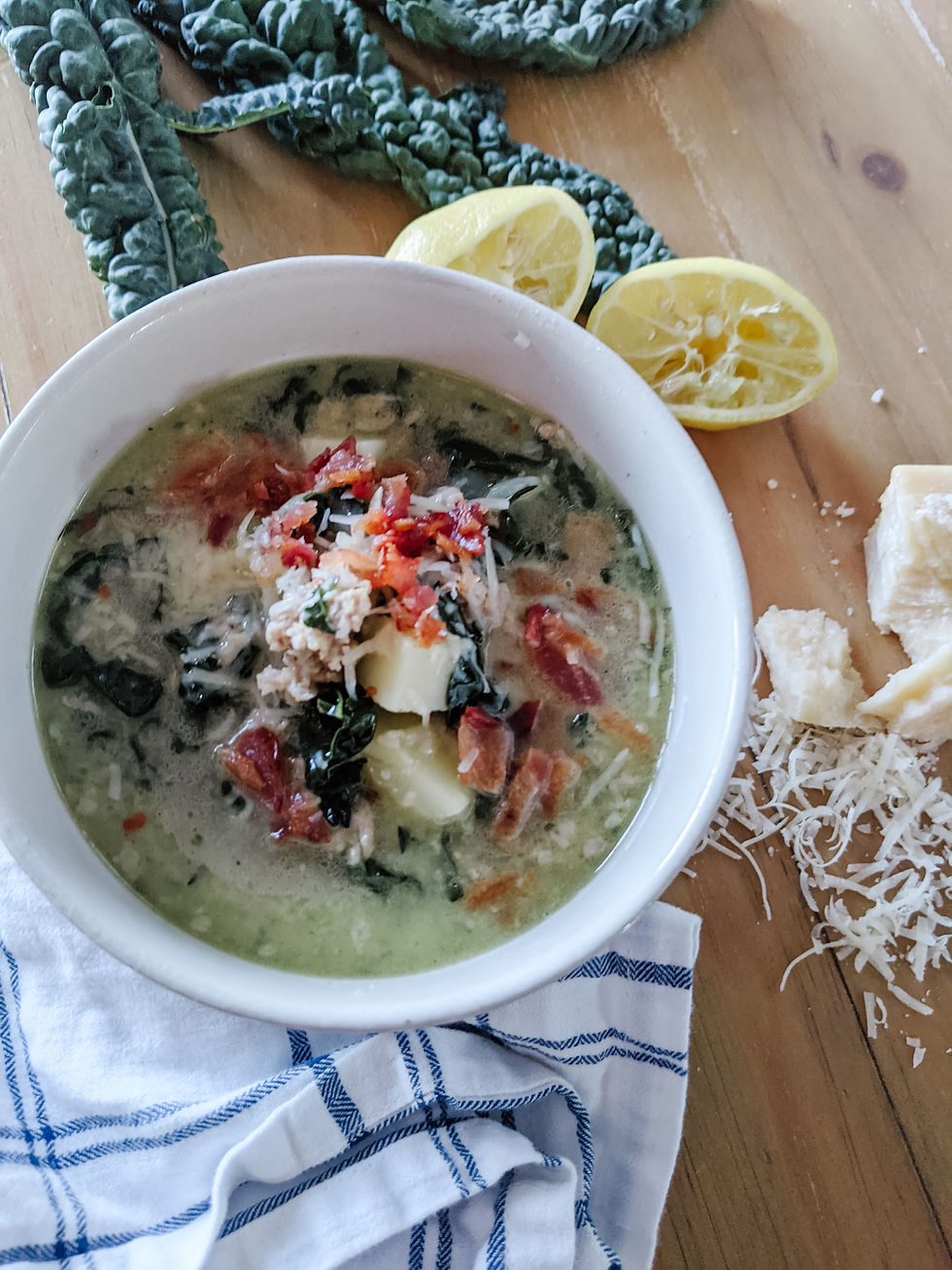 a Cozy bowl of dairy-free Zuppa Toscana, filled with cubed potatoes, bright green kale, ground sausage, and topped with crumbled bacon and parmesan cheese. The big, white soup bowl is surrounded by leaves of kale and juiced lemons