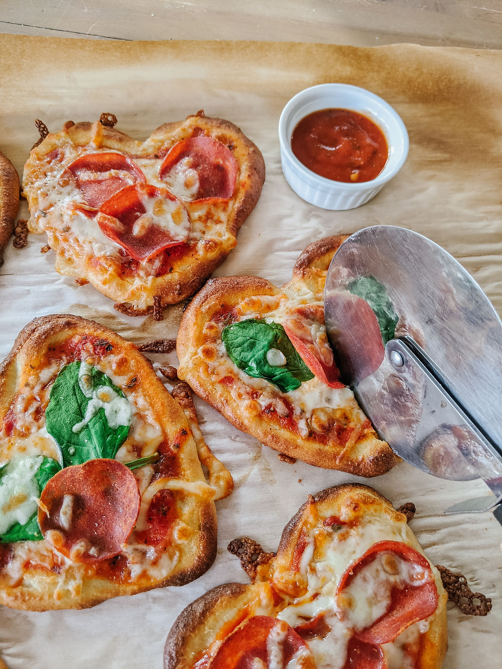 a cluster of four, heart-shaped, grain-free mini pizzas are shown. All contain various combinations of bubbly cheese, spinach, and pepperoni. One of the pizza's is getting cut in half with a pizza wheel. There's a small dish of marinara sauce to the top right corner.